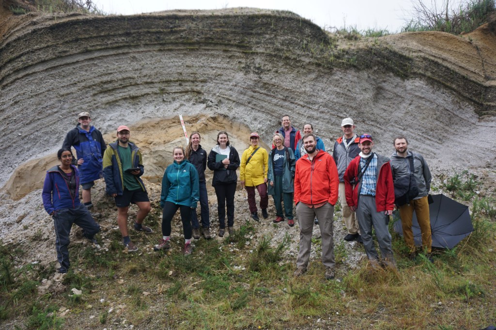 Tephra Seismites group members Tehnuka, Josh, and Richard (left) and workshop attendees, dressed in wet weather gear, stand in front of a white-grey and yellow pumice outcrop.